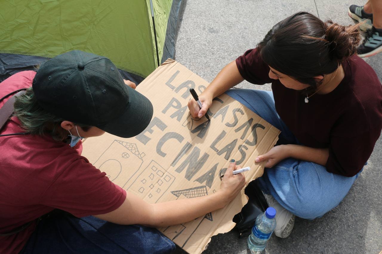 Dos personas pintan una pancarta de cartón con el lema «Las casas para las vecinas» en la acampada de Sol