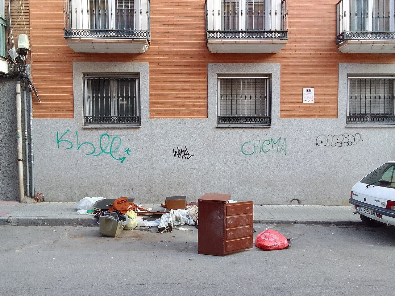Bolsas y residuos acumulados en la acera junto a una fachada de ladrillo con pintadas en la calle Serranillos