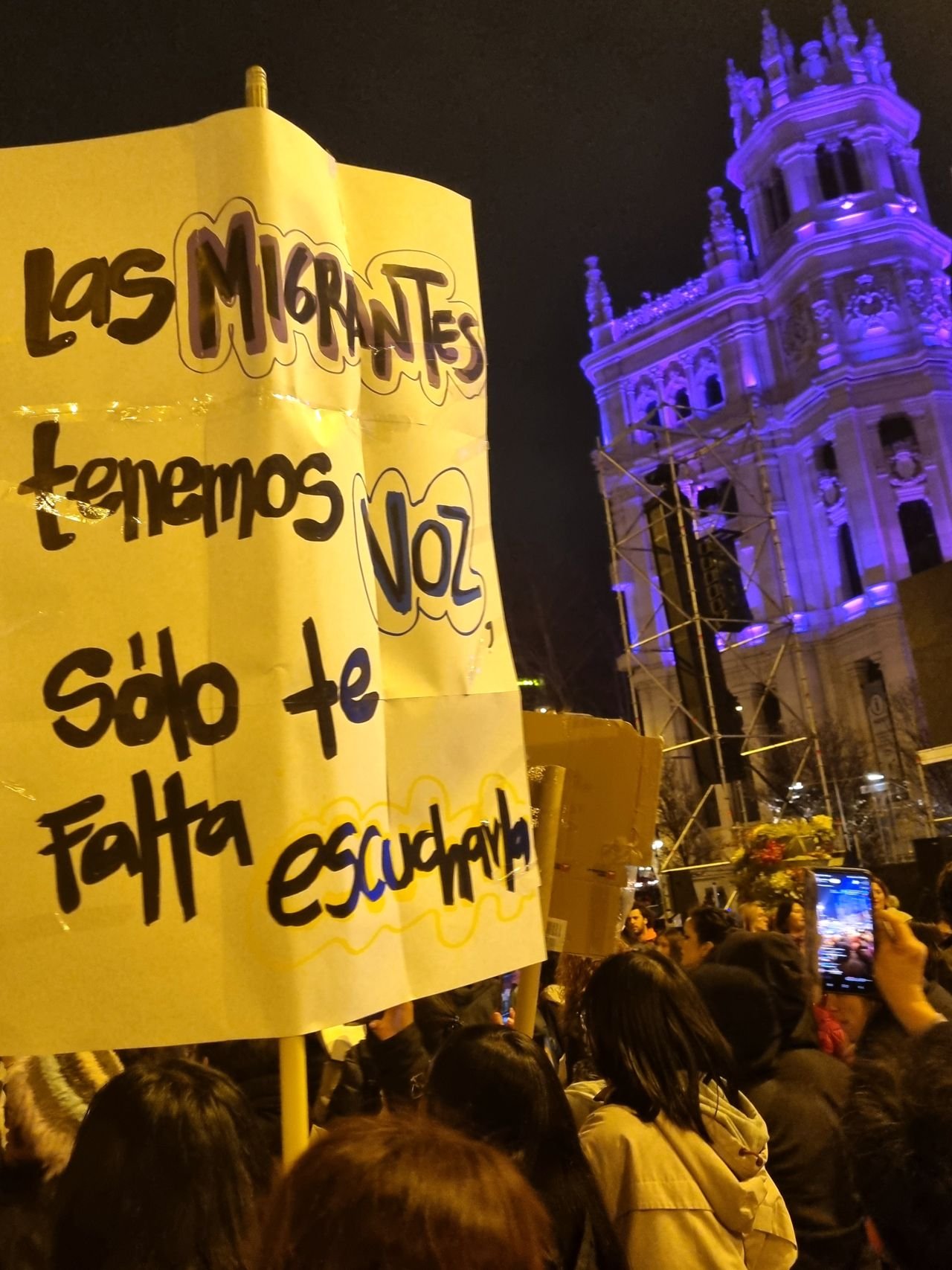 Cartel amarillo escrito a mano: «Las migrantes tenemos voz, sólo te falta escucharla», de noche frente al Palacio de Cibeles